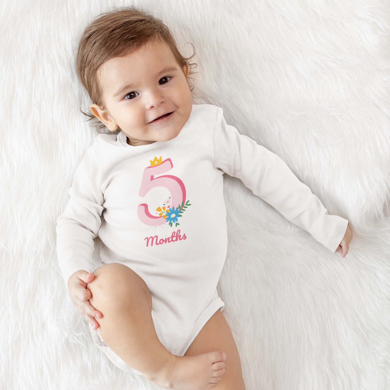 Baby wearing a white onesie with '5 Months' printed on it, lying on a white fluffy surface.