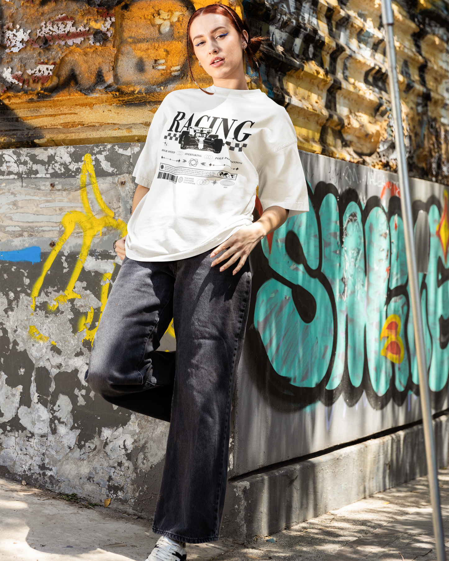 Person wearing a white t-shirt with racing text and graphics, leaning against a graffiti-covered wall.